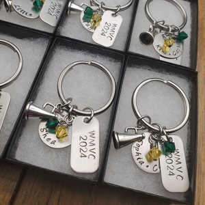 May include: Keychains with silver key rings, silver charms, and silver tags. The tags are engraved with the text "WMVC 2024" and names like "Sophia" and "Shea". The charms include a silver megaphone and green and yellow beads.