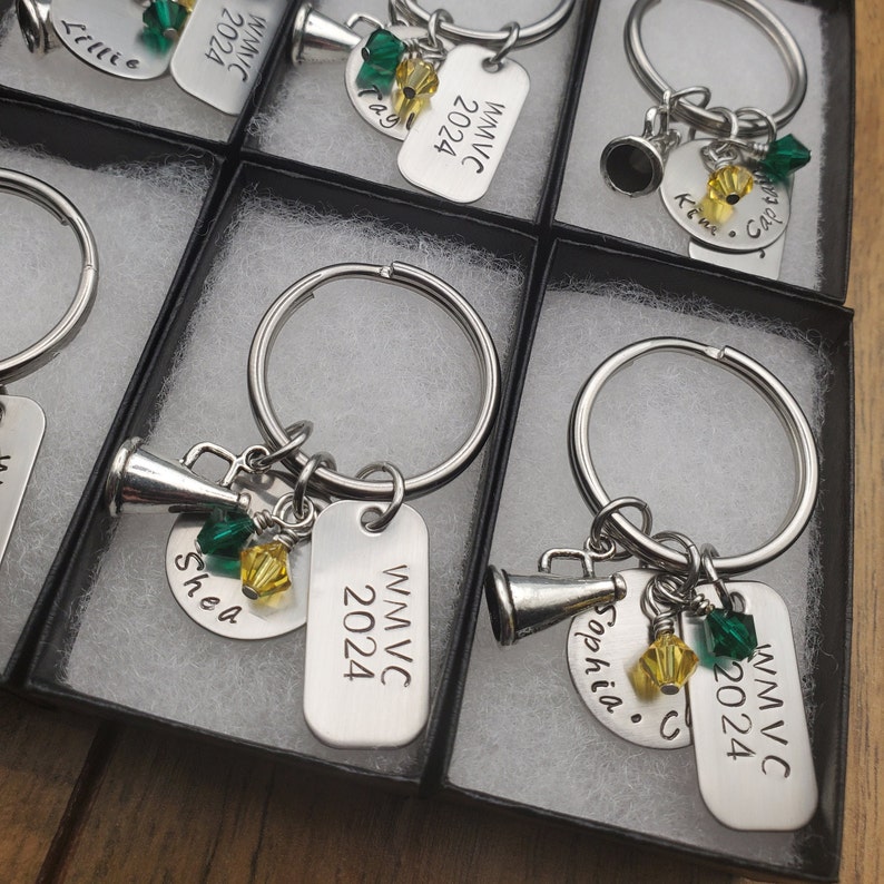 May include: Keychains with silver key rings, silver charms, and silver tags. The tags are engraved with the text "WMVC 2024" and names like "Sophia" and "Shea". The charms include a silver megaphone and green and yellow beads.