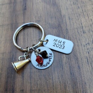 May include: A silver keychain with a silver charm that says "Annabella" and a silver charm that says "MHS 2023". The keychain also has a silver megaphone charm and red and black beads.