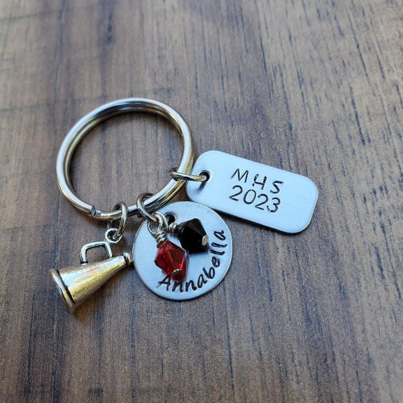 May include: A silver keychain with a silver charm that says "Annabella" and a silver charm that says "MHS 2023". The keychain also has a silver megaphone charm and red and black beads.