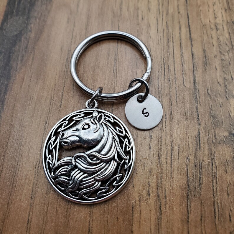 May include: Silver-toned horse keychain with a circular pendant featuring a detailed horse head design within a Celtic knot border. Includes a small, round tag with the letter "S". The keychain is attached to a silver key ring.