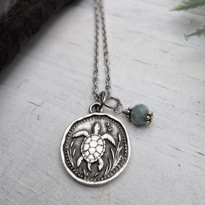Peut inclure: Un collier argenté avec un pendentif rond représentant une tortue. Le pendentif est suspendu à une fine chaîne, accompagné d'une petite perle facettée gris-vert. Ce collier d'inspiration nature est un bijou délicat.