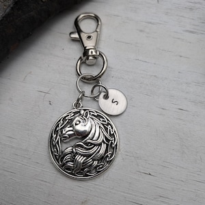 May include: Silver-toned keychain featuring a horse head pendant within a Celtic knot design. Includes a small, round charm with the letter "S". The keychain has a lobster clasp and key rings.