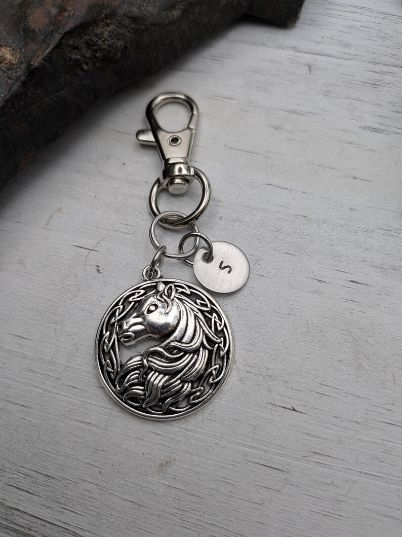 May include: Silver-toned keychain featuring a horse head pendant within a Celtic knot design. Includes a small, round charm with the letter "S". The keychain has a lobster clasp and key rings.