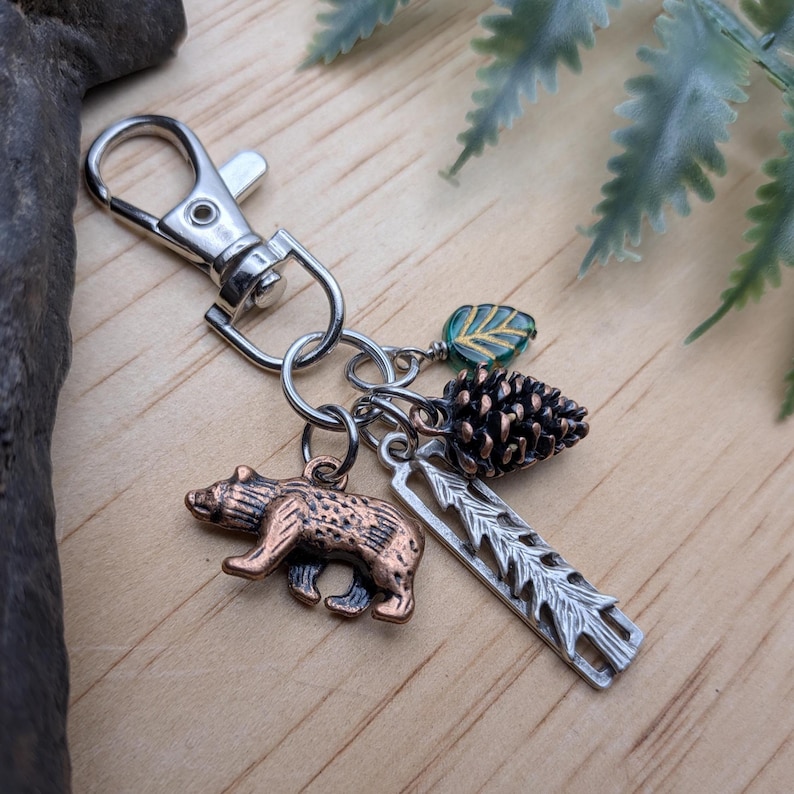 May include: A silver-tone keychain with a lobster clasp, featuring charms of a bear, pine cone, leaf, and a tree branch. The bear charm is bronze-colored. The pine cone is brown. The leaf is green and gold.