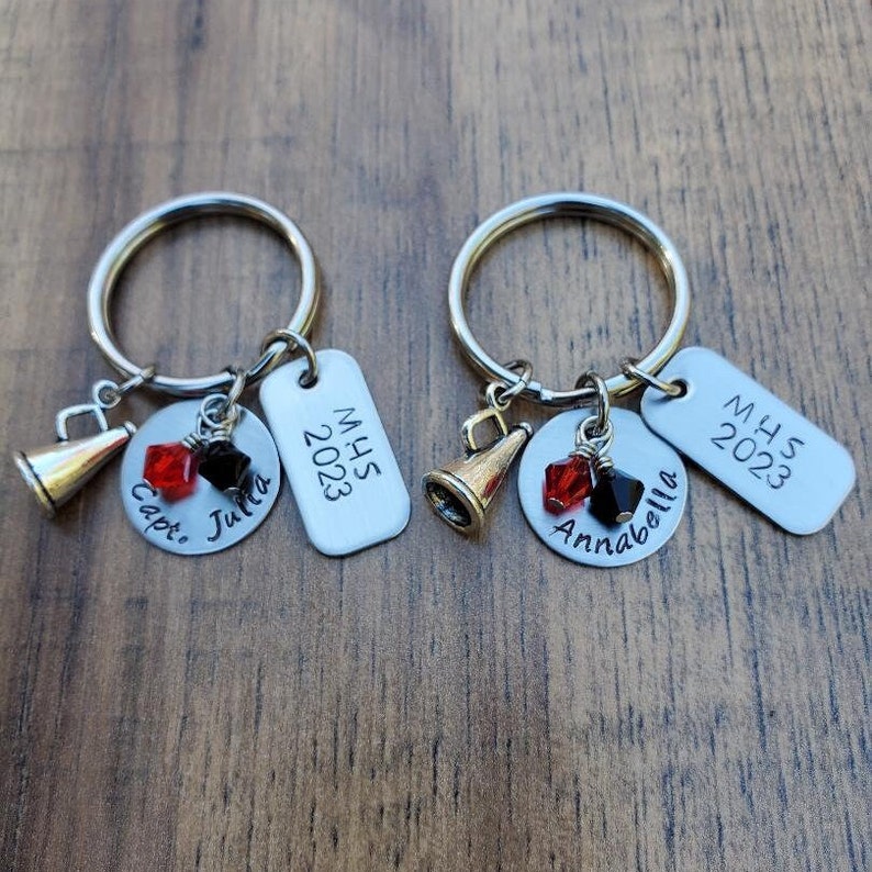 May include: Two silver keychains with charms. One keychain has a silver tag with the text "MHS 2023" and a charm with the text "Capt. Julia". The other keychain has a silver tag with the text "MHS 2023" and a charm with the text "Annabella".