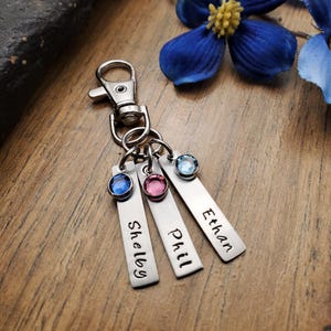 May include: A silver keychain with three rectangular tags, each engraved with a name: Shelby, Phil, and Ethan. Each tag has a colored gemstone: blue, pink, and light blue. The keychain has a clasp for easy attachment.