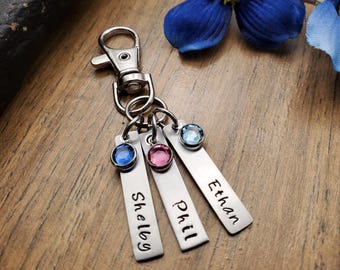 May include: A silver keychain with three rectangular tags, each engraved with a name: Shelby, Phil, and Ethan. Each tag has a colored gemstone: blue, pink, and light blue. The keychain has a clasp for easy attachment.