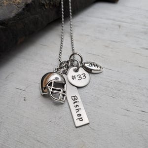 May include: A silver-toned necklace featuring a ball chain and three charms: a miniature football helmet, a round disc engraved with #33, and a rectangular bar pendant stamped with the name Bishop, alongside a small football charm.
