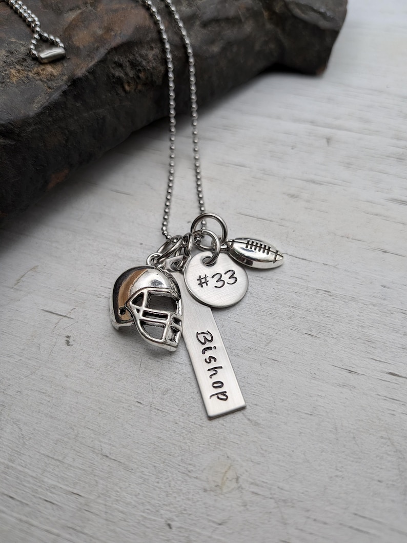 May include: A silver-toned necklace featuring a ball chain and three charms: a miniature football helmet, a round disc engraved with #33, and a rectangular bar pendant stamped with the name Bishop, alongside a small football charm.