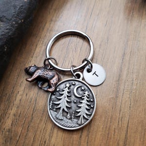 May include: A silver keychain with a bear charm and a circular pendant featuring trees, a crescent moon, and stars. Includes a small disc with the letter "T". The bear charm is bronze-colored.