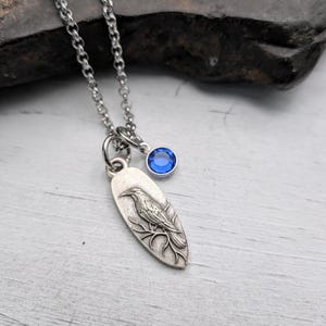 May include: Silver-toned necklace featuring a pendant with a detailed raven design perched on a branch. A small, round, blue gemstone charm hangs near the pendant. The chain is a delicate link style.