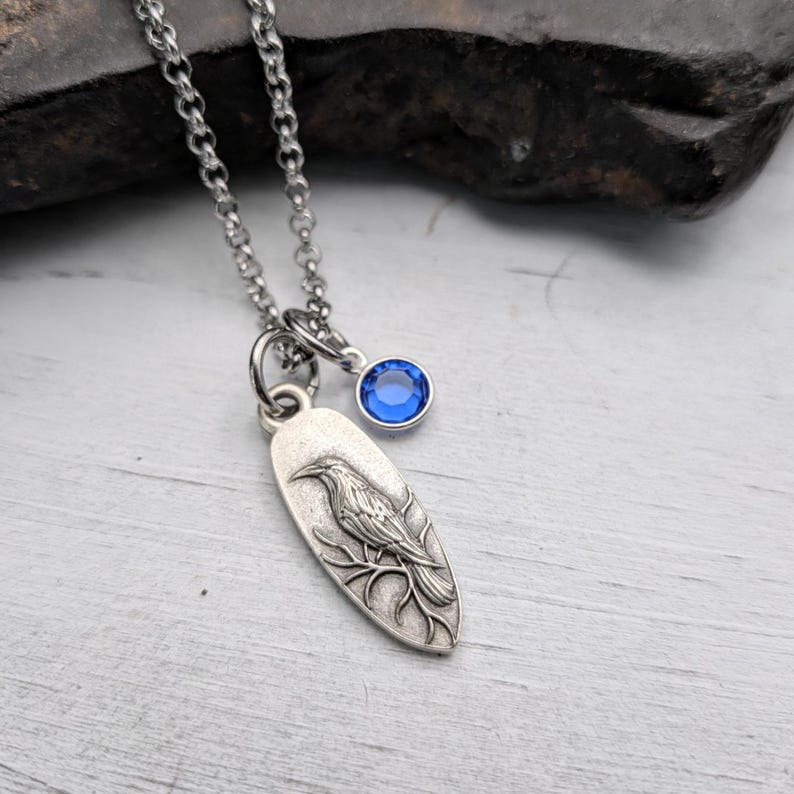 May include: Silver-toned necklace featuring a pendant with a detailed raven design perched on a branch. A small, round, blue gemstone charm hangs near the pendant. The chain is a delicate link style.