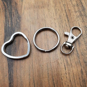 Puede incluir: Un conjunto de accesorios para llaveros de color plateado, que incluye un anillo en forma de coraz&oacute;n, un anillo redondo y un mosquet&oacute;n. Los art&iacute;culos se muestran sobre una superficie de madera.