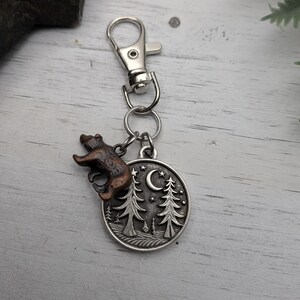 May include: A silver-colored keychain with a bear charm and a circular pendant featuring a forest scene with trees, a crescent moon, and stars. The bear charm is brown and bronze. The keychain has a clasp for easy attachment.