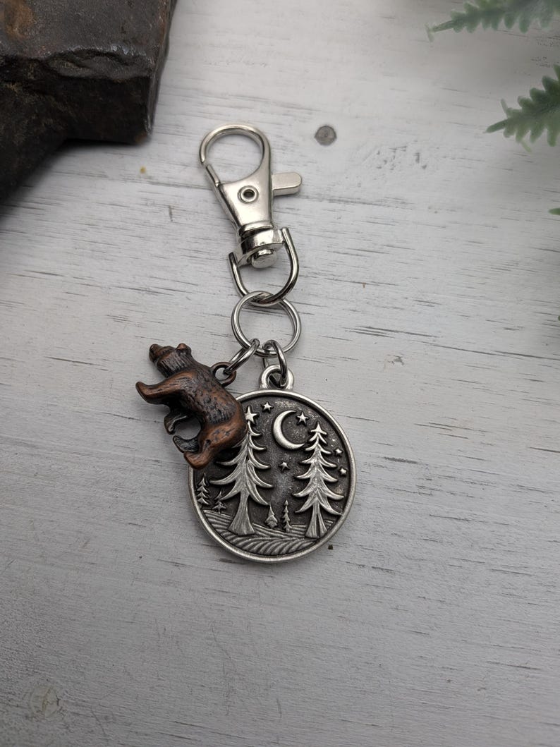 May include: A silver-colored keychain with a bear charm and a circular pendant featuring a forest scene with trees, a crescent moon, and stars. The bear charm is brown and bronze. The keychain has a clasp for easy attachment.