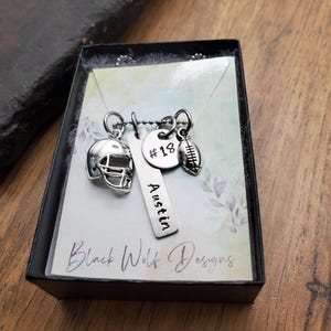 May include: A silver-toned necklace featuring three charms: a football helmet, a vertical rectangular bar engraved with the name Austin, and a round disc stamped with #18, plus a small football charm, presented on a display card in a black box.