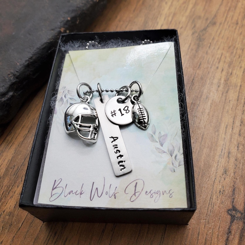 May include: A silver-toned necklace featuring three charms: a football helmet, a vertical rectangular bar engraved with the name Austin, and a round disc stamped with #18, plus a small football charm, presented on a display card in a black box.
