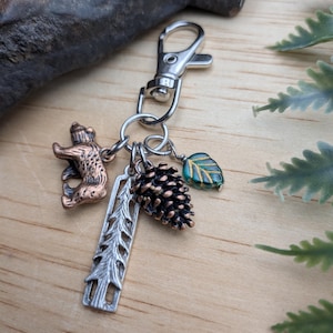 May include: A silver-toned keychain with multiple charms. Charms include a bear, a pine cone, a leaf, and a tree-shaped charm. The bear is bronze-colored, the leaf is teal and gold, and the pine cone is brown. The keychain is on a wooden surface.