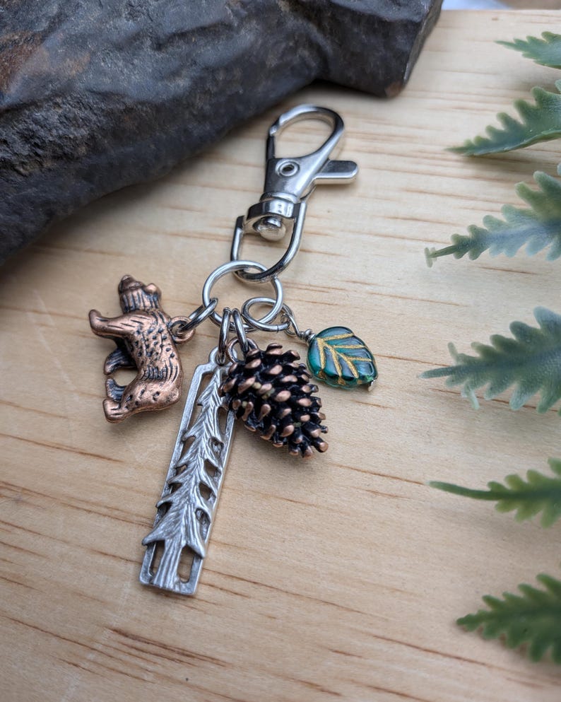May include: A silver-toned keychain with multiple charms. Charms include a bear, a pine cone, a leaf, and a tree-shaped charm. The bear is bronze-colored, the leaf is teal and gold, and the pine cone is brown. The keychain is on a wooden surface.