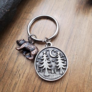 May include: A silver-toned keychain with two charms. One charm is a detailed bear, the other is a round pendant with a forest scene, a crescent moon, and stars. The keychain is on a wooden surface.