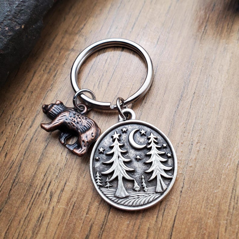 May include: A silver-toned keychain with two charms. One charm is a detailed bear, the other is a round pendant with a forest scene, a crescent moon, and stars. The keychain is on a wooden surface.