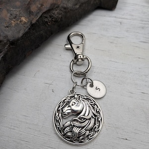 May include: Silver-toned keychain featuring a detailed horse head pendant within a Celtic knot design. Includes a small, round charm with the letter "S". The keychain has a lobster clasp for easy attachment.