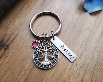May include: Silver keychain with a Libra zodiac symbol charm, a pink crystal charm, and a personalized tag that reads "Ashley".