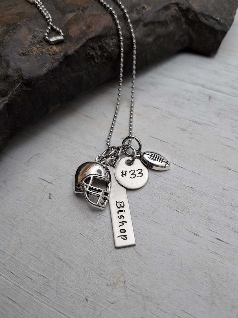 May include: A silver-toned ball chain necklace featuring three charms: a miniature football helmet, a round disc engraved with #33, and a rectangular vertical bar tag engraved with the name Bishop, accompanied by a small football charm.