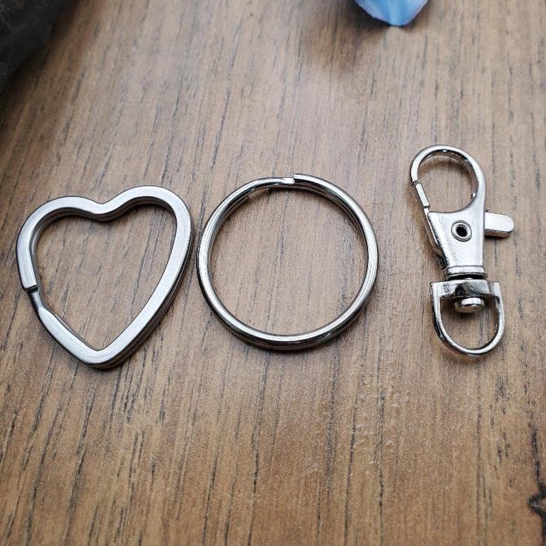 May include: A collection of silver-colored keychains and clasps. The set includes a heart-shaped ring, a circular ring, and a lobster clasp. The items are displayed on a wooden surface.