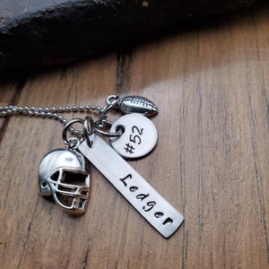May include: A silver-toned necklace featuring a ball chain and three charms: a miniature football helmet, a small round disc stamped with #52, and a rectangular metal bar engraved with the name Ledger in a simple black sans-serif font.