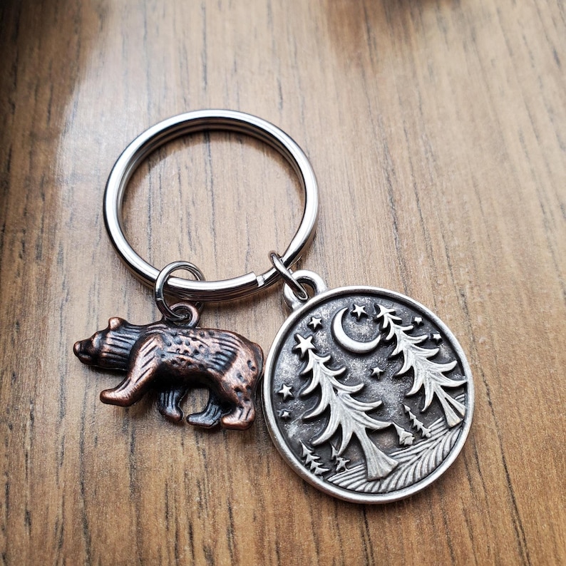 May include: A silver-colored keychain with two charms. One charm is a small, bronze-colored bear. The other is a circular charm with a forest scene, a crescent moon, and stars. The keychain is on a wooden surface.