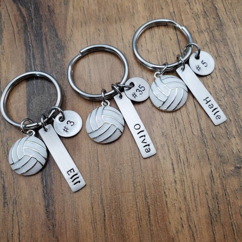 May include: Three silver volleyball keychain charms with a white volleyball charm, a silver charm with a number and a silver bar charm with a name. The names are Ellie, Olivia, and Halle.