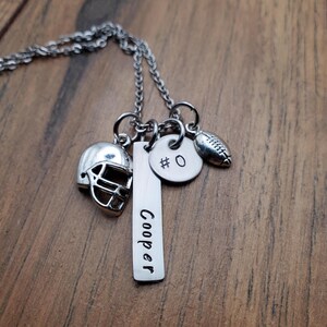 May include: A silver-toned metal necklace featuring three charms: a football helmet, a rectangular bar pendant engraved with the name Cooper, and a circular disc pendant stamped with the text #0, alongside a small football-shaped charm.