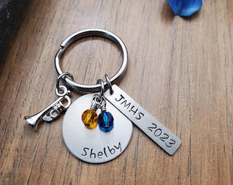 May include: A silver keychain with a trumpet charm, blue and yellow beads, and two tags. The larger tag says "Shelby" and the smaller tag says "JMHS 2023".