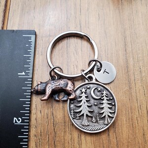 May include: A metal keychain with a bear charm and a round pendant featuring trees, a crescent moon, and stars. The keychain includes a small initial charm. The ruler shows the keychain is approximately 2 inches long.