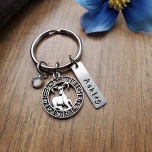 May include: Silver keychain with a zodiac symbol charm, a crystal charm, and a personalized tag that says "Ashley".