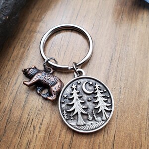 May include: A silver-colored keychain with two charms. One charm is a small, bronze-colored bear. The other is a round charm with a forest scene, including trees, a crescent moon, and stars. The keychain is on a wooden surface.