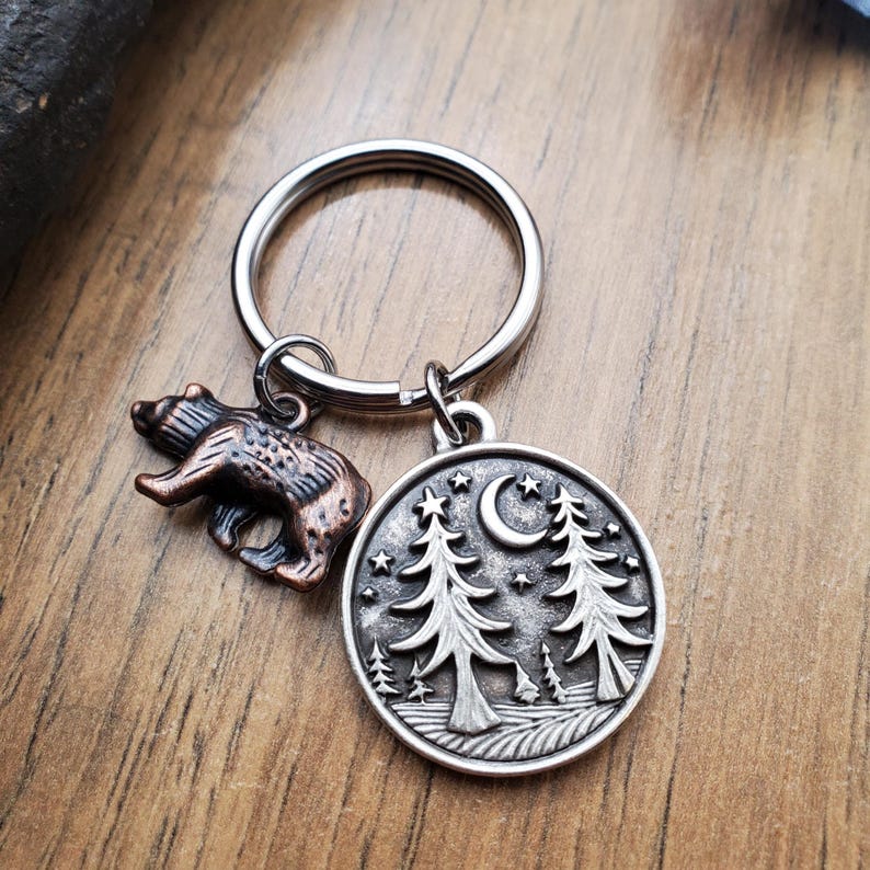 May include: A silver-colored keychain with two charms. One charm is a small, bronze-colored bear. The other is a round charm with a forest scene, including trees, a crescent moon, and stars. The keychain is on a wooden surface.