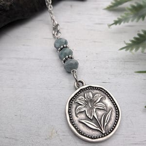 May include: A silver necklace featuring a pendant with a lily flower design. The pendant is round with a beaded border. The necklace includes a chain and several light blue beads.