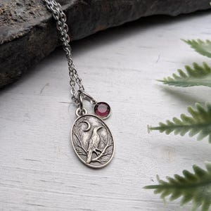 May include: Silver-toned pendant necklace featuring an oval pendant with a detailed raven design, a crescent moon, and a small, round, red gemstone. The necklace has a delicate chain and is set against a white wooden surface.