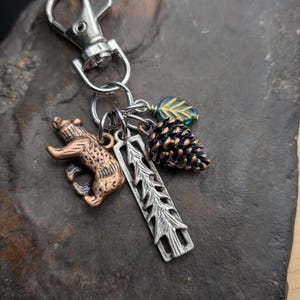 May include: A silver-tone keychain with a lobster clasp, featuring charms of a bear, pine cone, leaf, and a tree-shaped pendant. The bear charm has a bronze finish, and the pine cone is brown. The leaf charm is green.