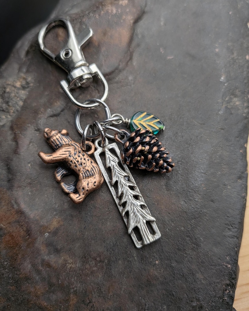 May include: A silver-tone keychain with a lobster clasp, featuring charms of a bear, pine cone, leaf, and a tree-shaped pendant. The bear charm has a bronze finish, and the pine cone is brown. The leaf charm is green.