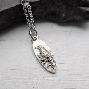 May include: Silver-toned raven pendant necklace. The oval pendant features a detailed raven perched on a branch. The necklace has a delicate chain and a secure clasp. The raven is intricately designed, showcasing feathers and a sharp beak.