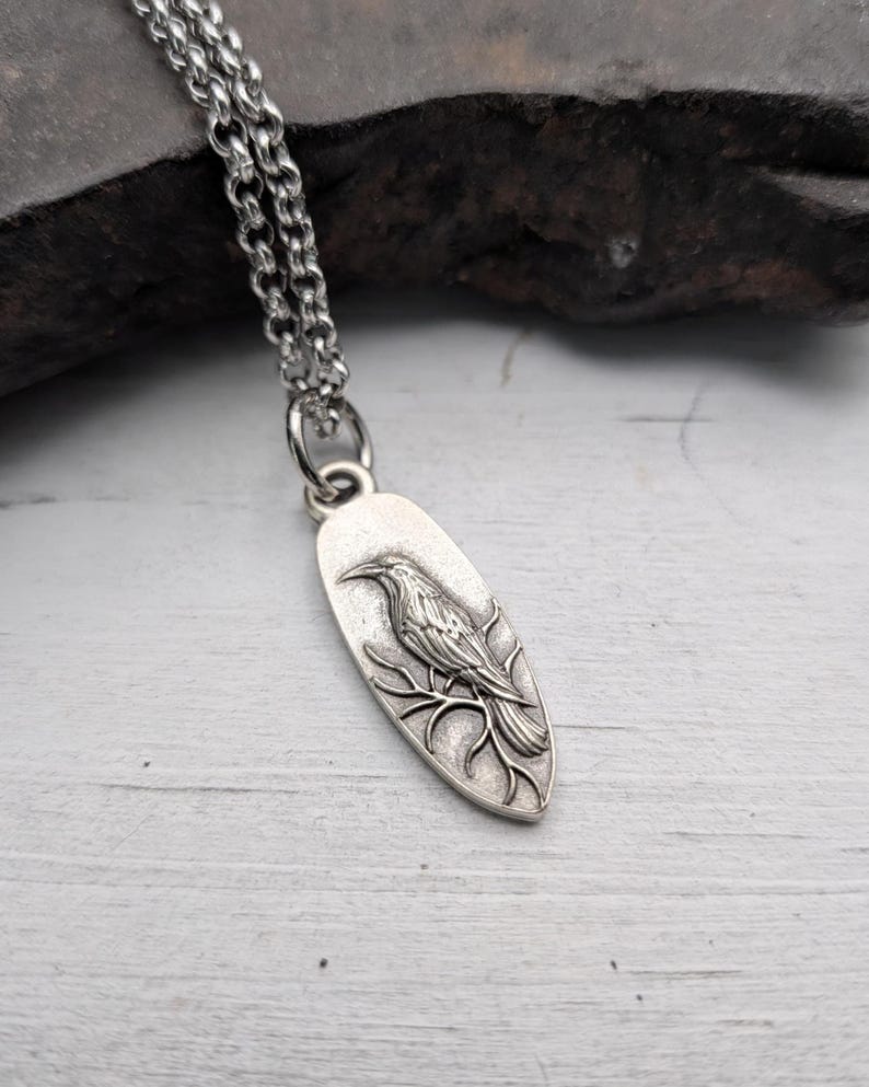 May include: Silver-toned raven pendant necklace. The oval pendant features a detailed raven perched on a branch. The necklace has a delicate chain and a secure clasp. The raven is intricately designed, showcasing feathers and a sharp beak.