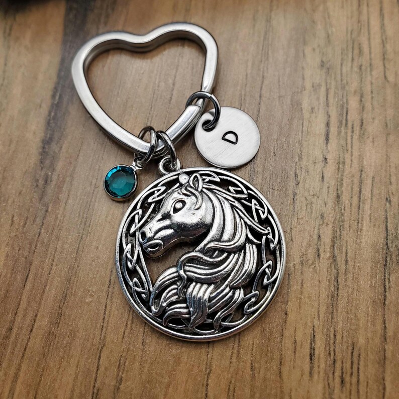 May include: Silver-toned keychain with a heart-shaped top and a round charm featuring a horse head design. Includes a teal-colored gemstone and a small disc with the letter "D". The horse charm is surrounded by an intricate Celtic knot design.