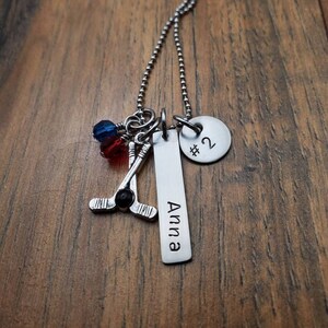 May include: Silver necklace with a hockey stick charm, a red and blue crystal charm, a round charm with the number "#2" and a rectangular charm with the name "Anna" engraved on it.