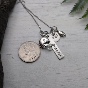 May include: A silver-toned necklace featuring a ball chain and multiple charms: a football helmet, a small football, a round disc stamped with #64, and a rectangular bar engraved with the name Bragden, shown next to a U.S. quarter coin.