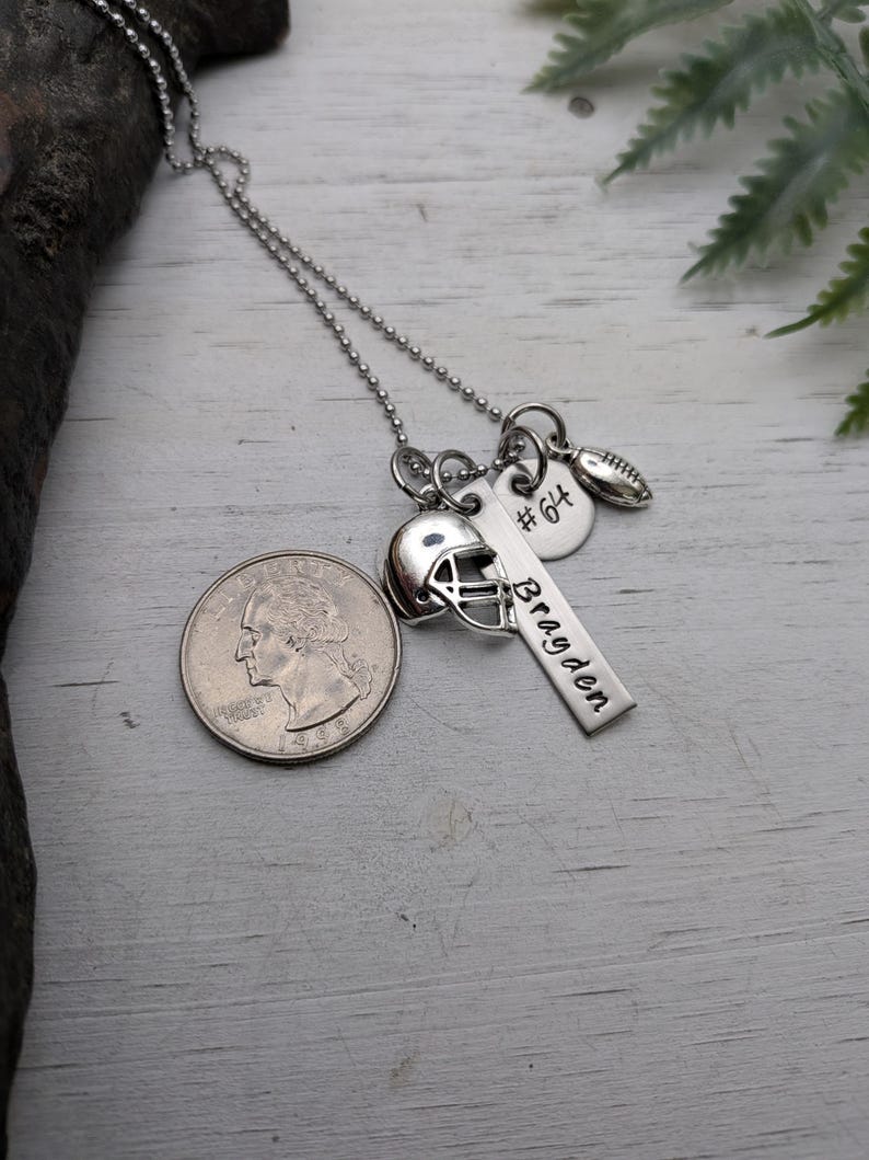 May include: A silver-toned necklace featuring a ball chain and multiple charms: a football helmet, a small football, a round disc stamped with #64, and a rectangular bar engraved with the name Bragden, shown next to a U.S. quarter coin.