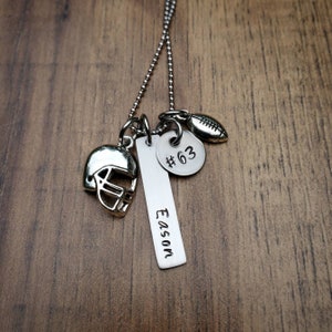 May include: A silver-toned necklace featuring a ball chain and three charms: a football helmet, a rectangular bar pendant engraved with the name Eason, and a circular disc pendant stamped with the text #63, alongside a small football charm.